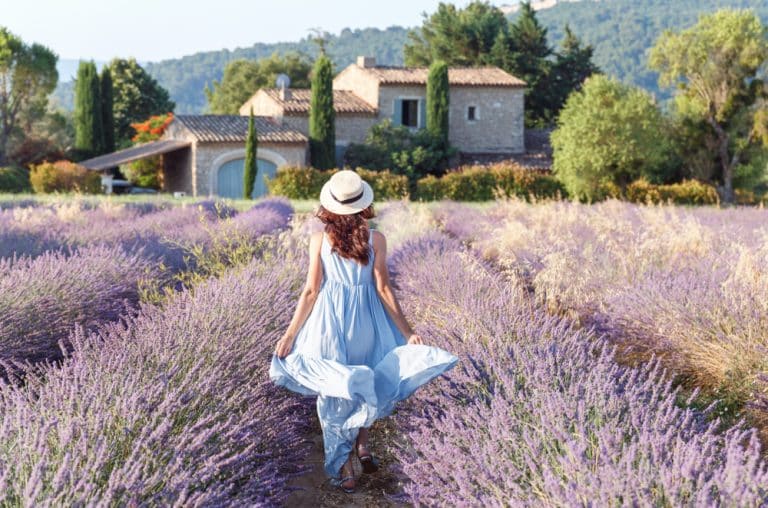 femme portant un chapeau marchant dans un champ de lavande avec une maison en arrière-plan