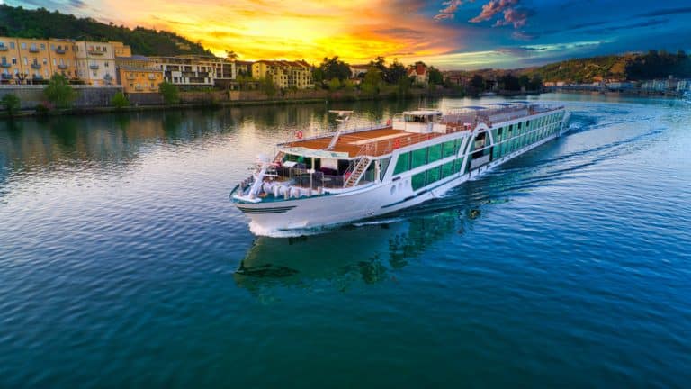 bateau de croisière naviguant sur un fleuve au coucher du soleil avec des bâtiments colorés en arrière-plan