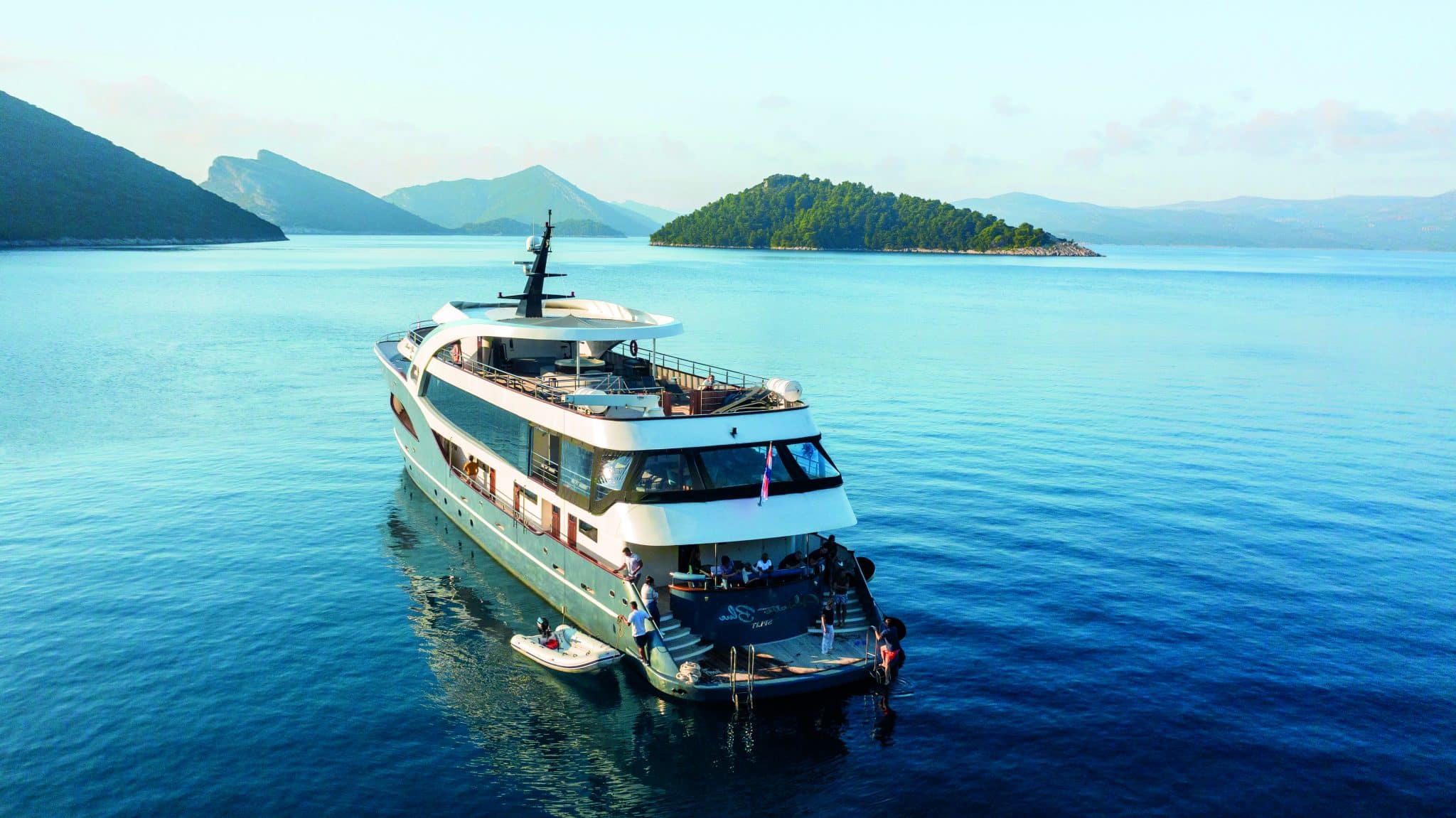 yacht de luxe blanc et bleu ancré en Méditerranée face à une île boisée et des montagnes