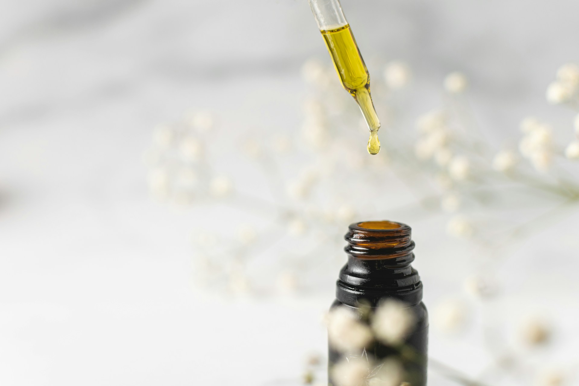 goutte d'huile cbd tombant d'un pipette dans un flacon ambré avec fleurs blanches