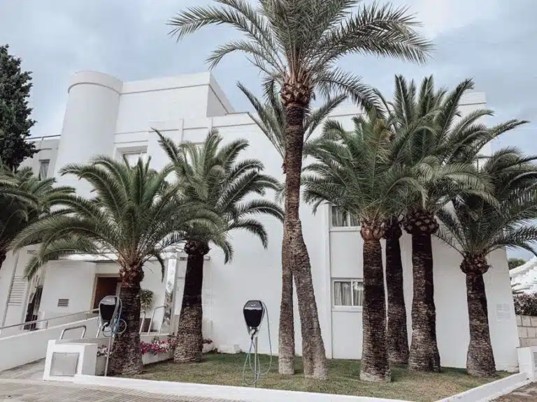 Contemporary white building facade with palm trees and charging stations on a city street.