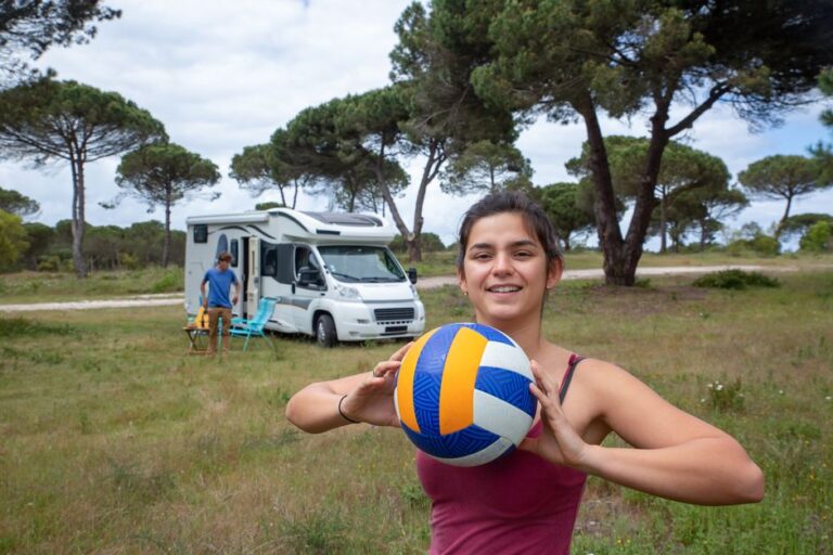 Smiling young woman holding volleyball near camper van in scenic Portugal campsite.